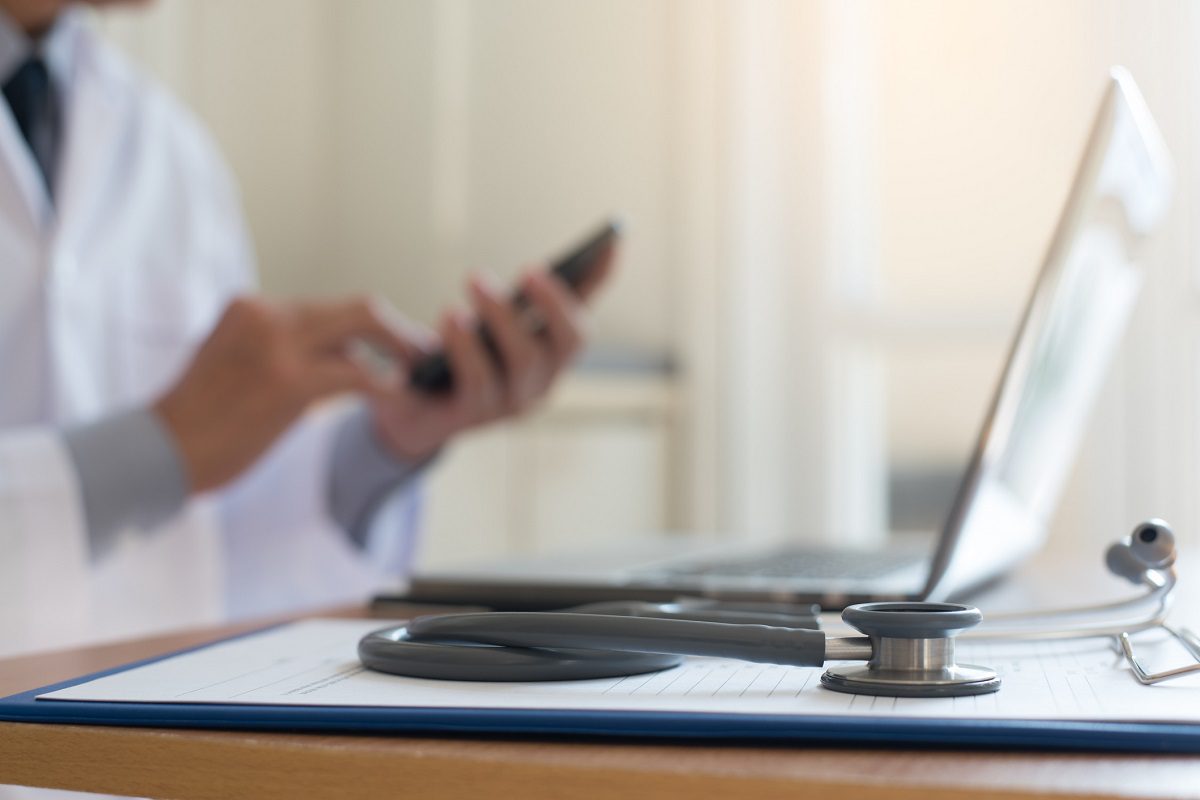 A stethoscope rests on a clipboard next to a laptop, while a person in a white coat uses a smartphone in the background.