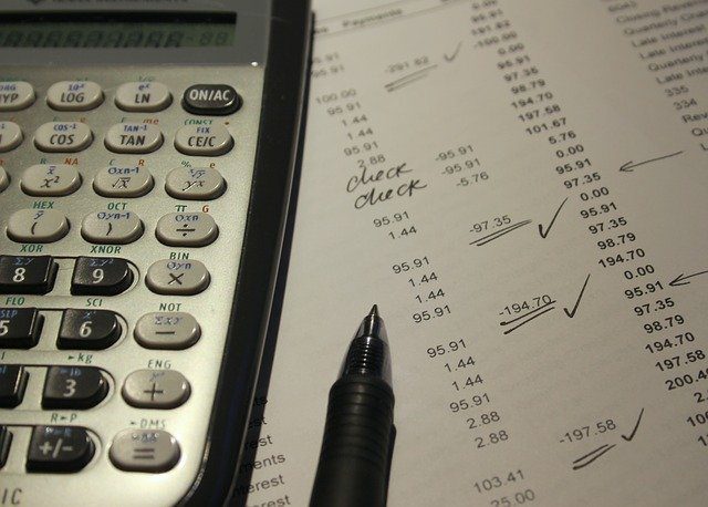 A calculator, a pen, and a printed list of numbers with several figures checked and handwritten notes that read check, suggesting financial review or accounting work.