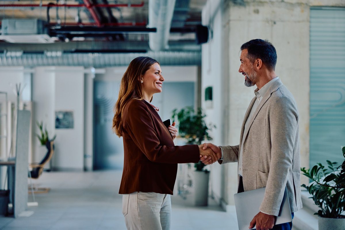 Two confident business professionals, a mid adult woman and man, smile while shaking hands in a modern office, symbolizing partnership, agreement, and professional success