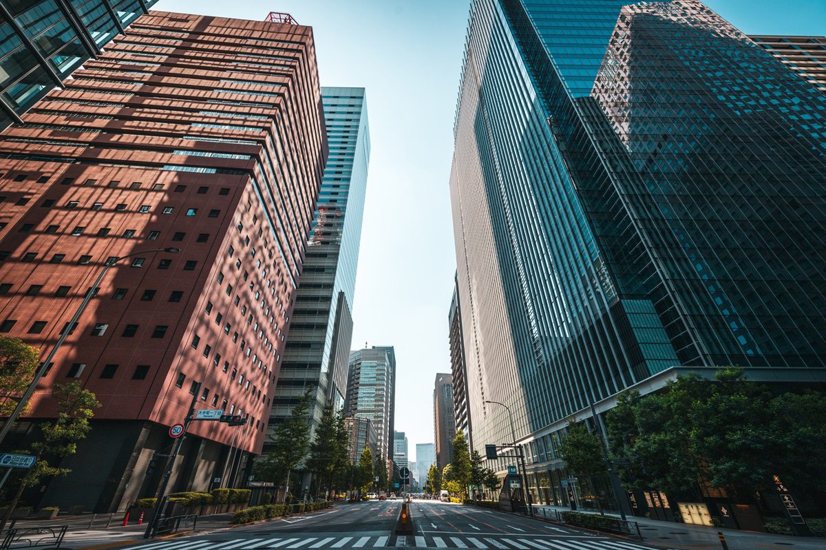 A wide city street flanked by tall modern office buildings on both sides under a clear blue sky. The street appears empty with no visible vehicles or pedestrians.