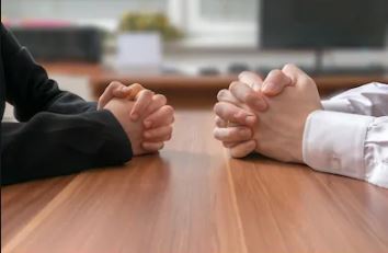 Two people sit across from each other at a wooden table with their hands clasped, suggesting a meeting or discussion.