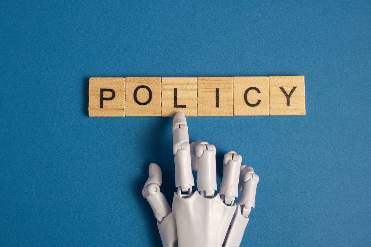 White robotic hand pointing to the word policy on wooden blocks over a blue background representing artificial intelligence ethics and government regulations.