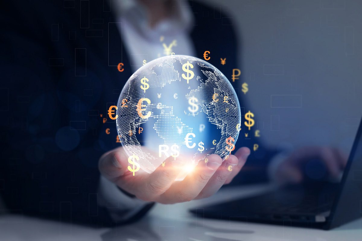 A person holding a glowing digital globe with currency symbols, including dollar, euro, and yen, in front of a laptop.