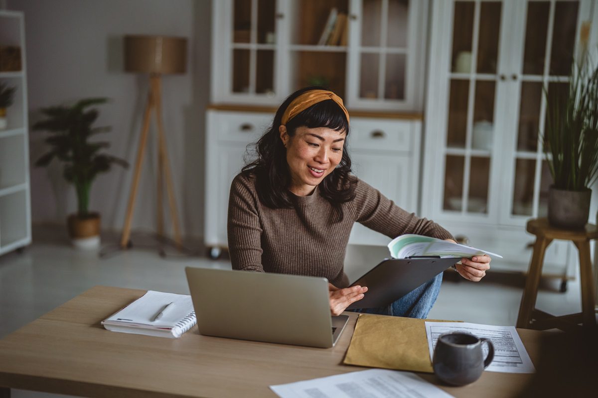 Asian woman smiling and concentrating while working remotely from her spacious home office, reviewing paperwork and using a laptop for productivity and business tasks