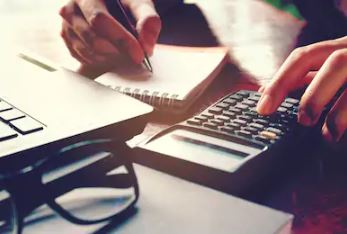 Person using a calculator and writing in a notebook beside a laptop and eyeglasses on a desk.