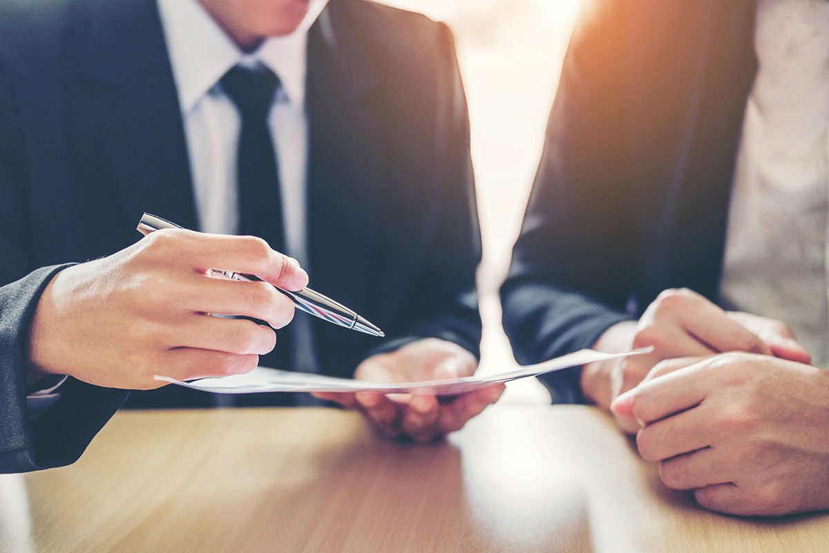 Two people in business attire sit at a table, one holding a pen and pointing to a document, while the other listens attentively.