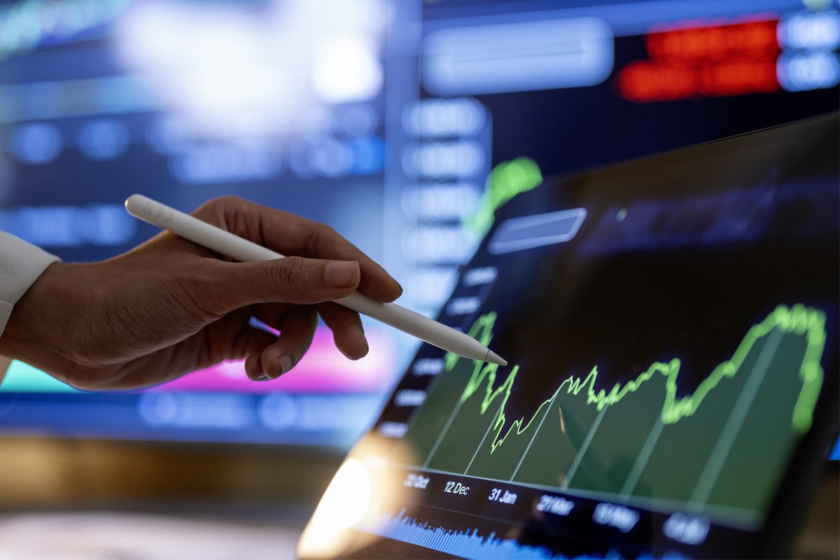 A person uses a stylus to analyze a fluctuating line graph on a digital tablet, with financial data and charts displayed on large screens in the background.