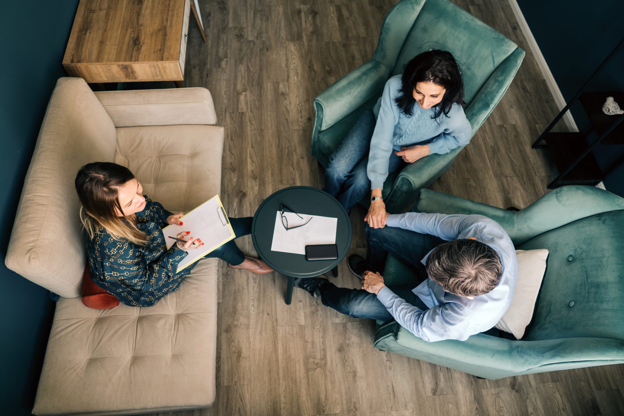 Three people sit in armchairs around a small table with papers, a pen, and a phone, engaged in conversation, viewed from above.