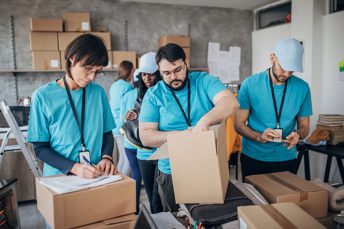 Three people in blue shirts and lanyards work together in a room, packing boxes and filling out paperwork, with shelves of packages in the background.