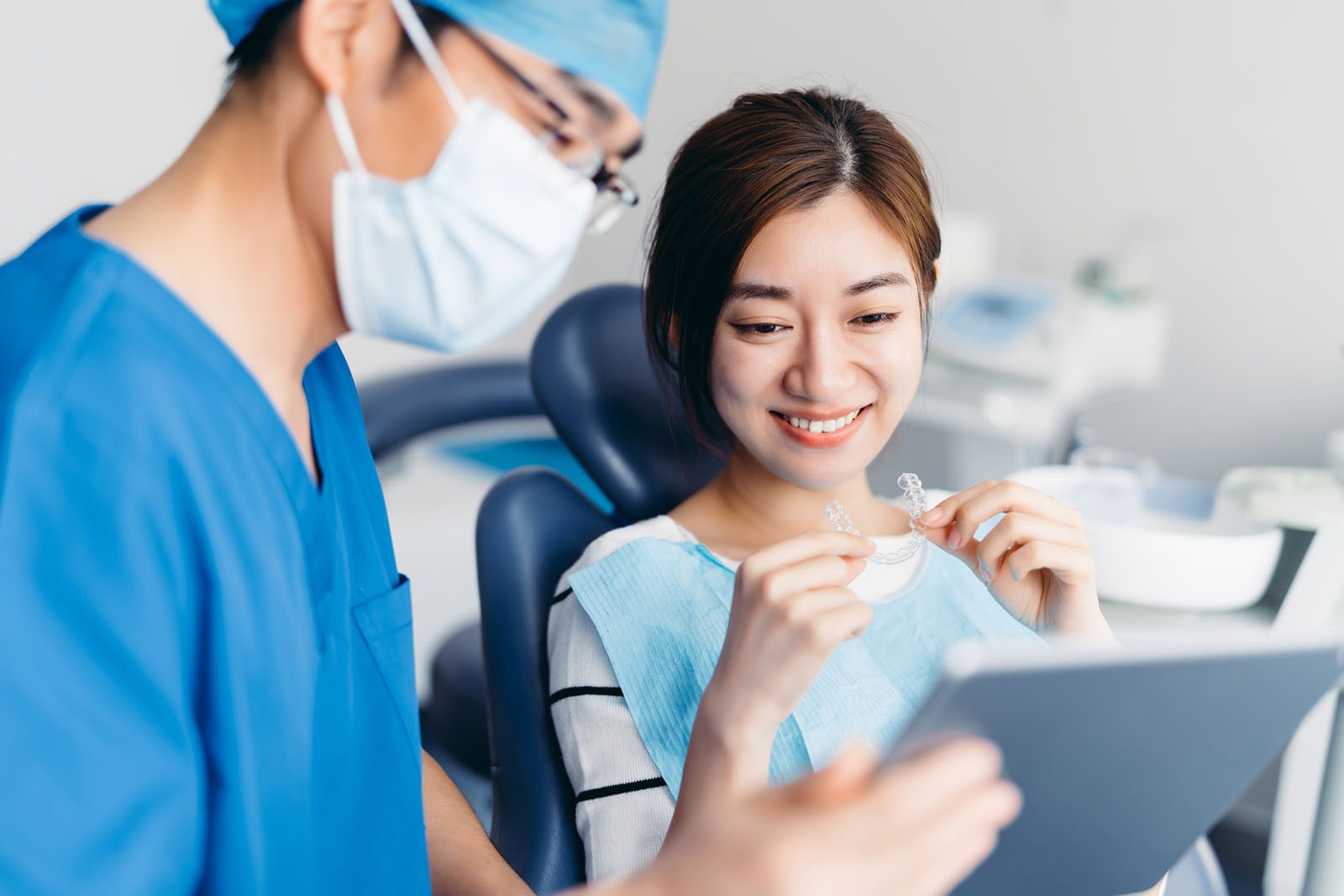 A dentist in scrubs and mask consults with a smiling patient holding clear aligners, both looking at a digital tablet in a dental clinic.