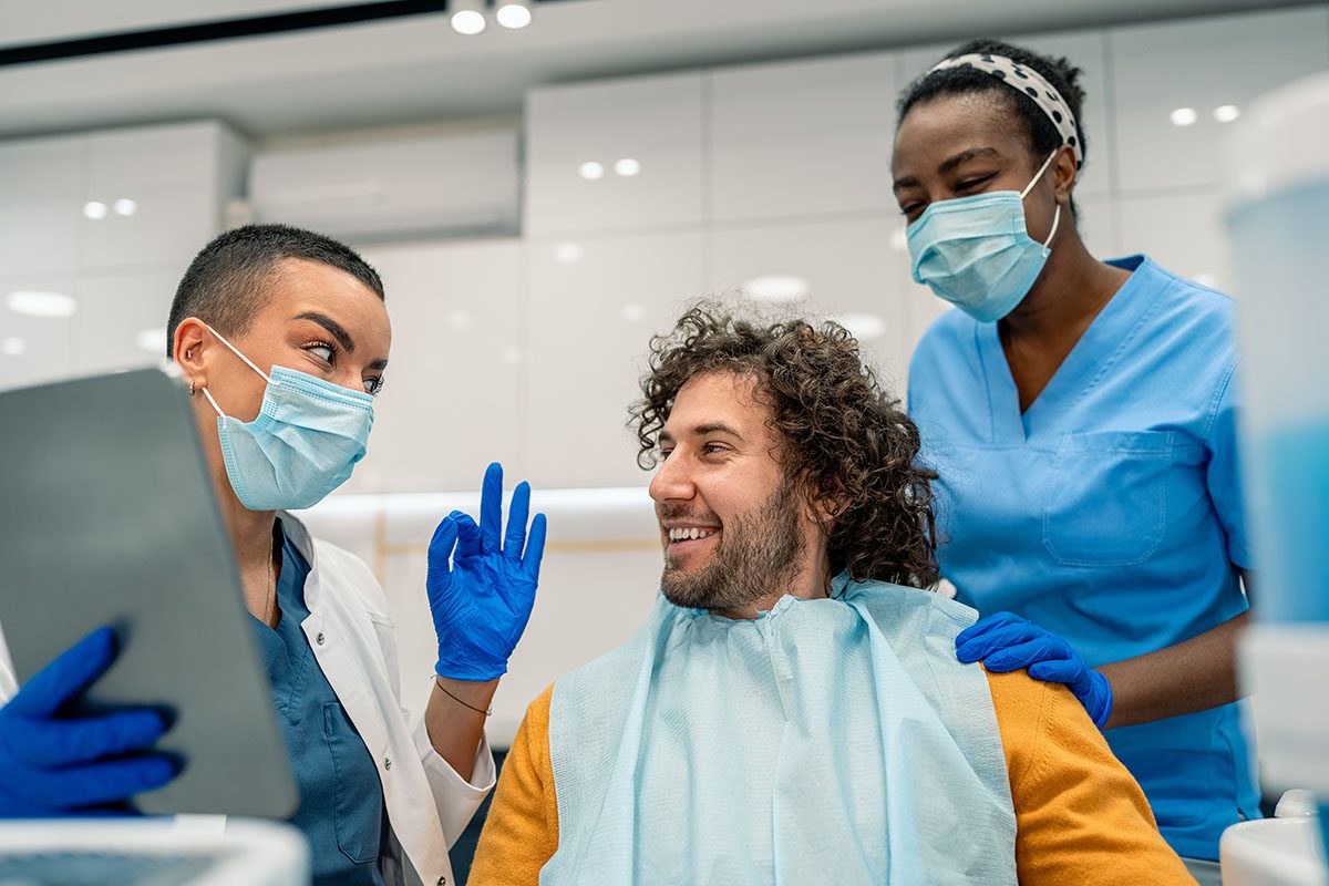 A patient in a dental chair smiles at two dental professionals wearing masks and gloves; one holds a tablet and gestures ok, while the other stands beside the patient.
