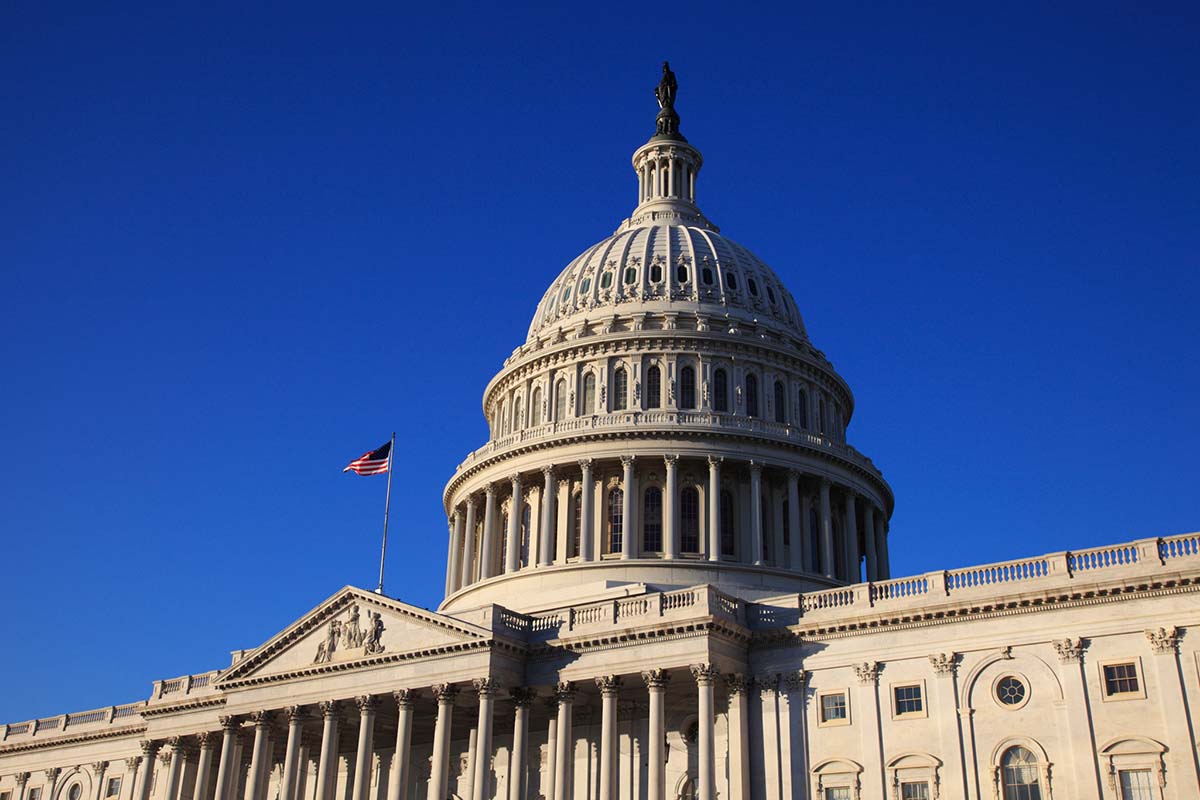 The United States Capitol building with the dome and an American flag visible against a clear blue sky.