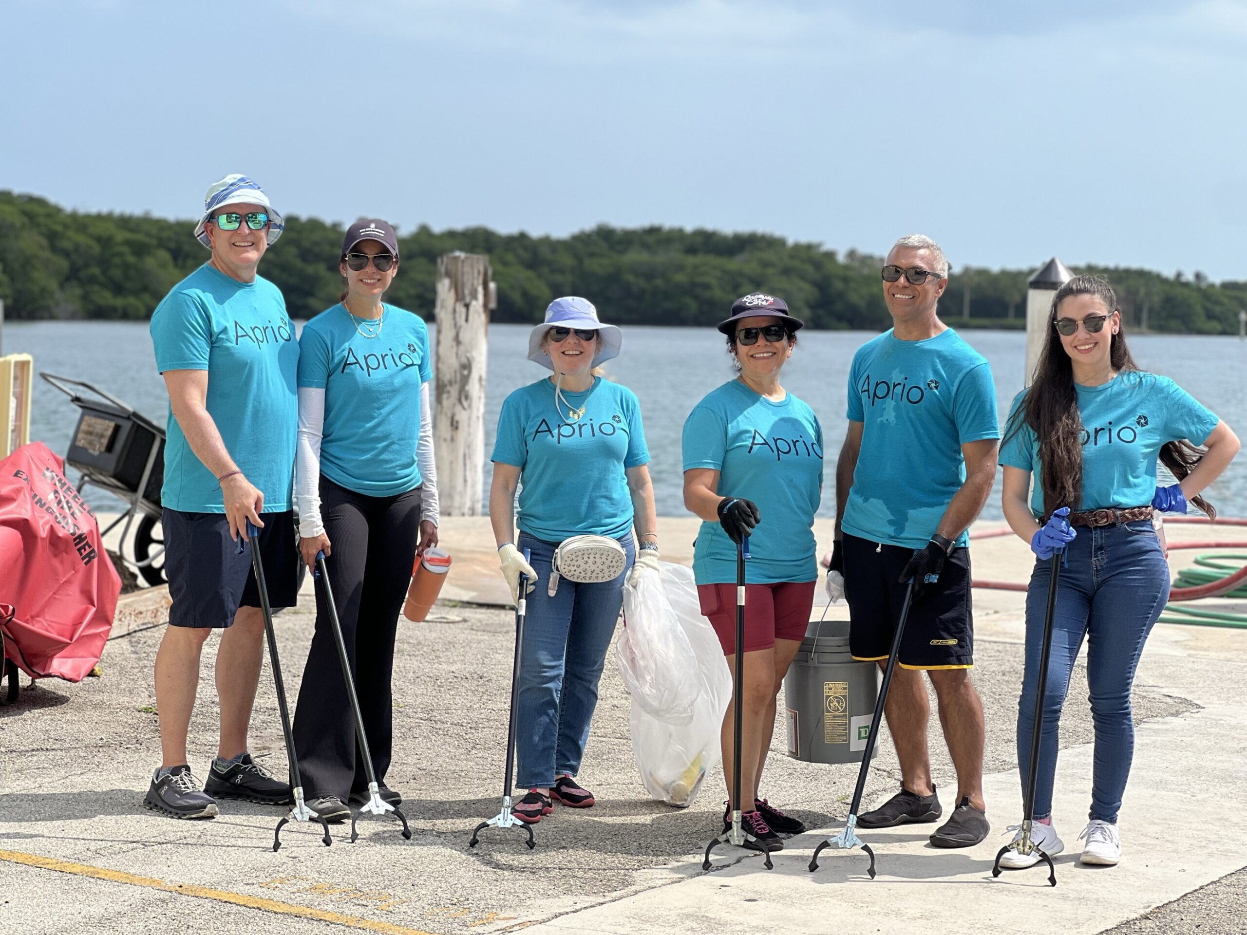 Six people wearing matching blue Aprio shirts stand on a waterfront holding litter pickers and trash bags, participating in a cleanup activity.