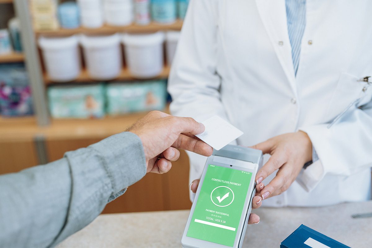 A person makes a contactless payment with a card on a digital device held by a pharmacist; the screen shows a payment confirmation.