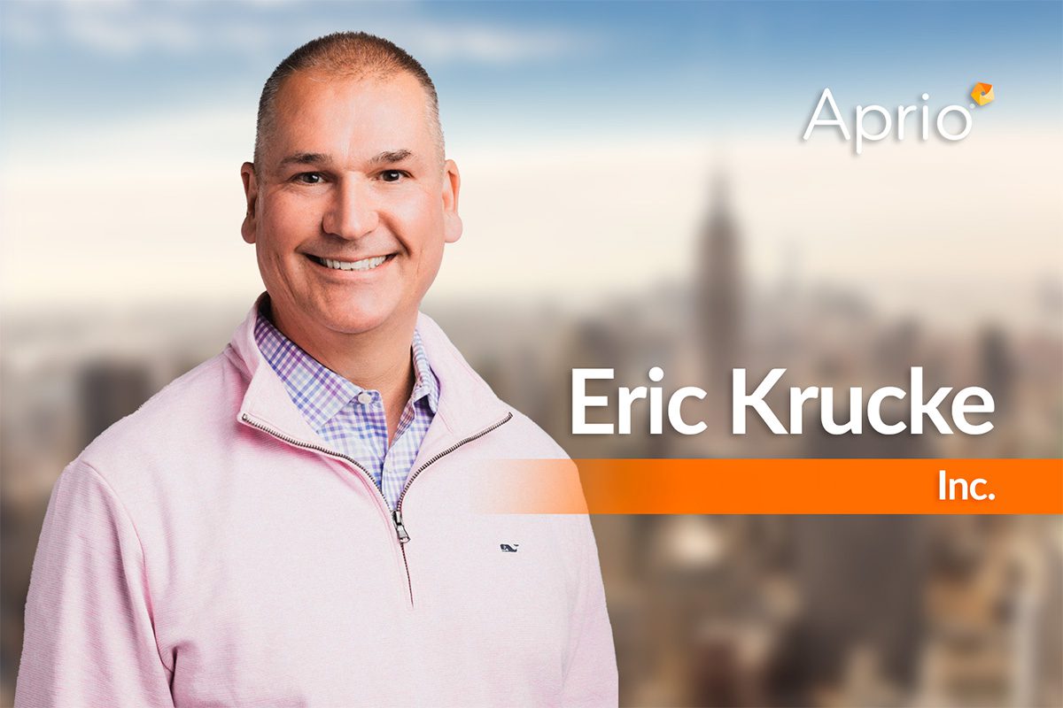 A man in a light pink sweater smiles at the camera with “Eric Krucke” and “Aprio Inc.” written beside him; the background is a blurred cityscape.