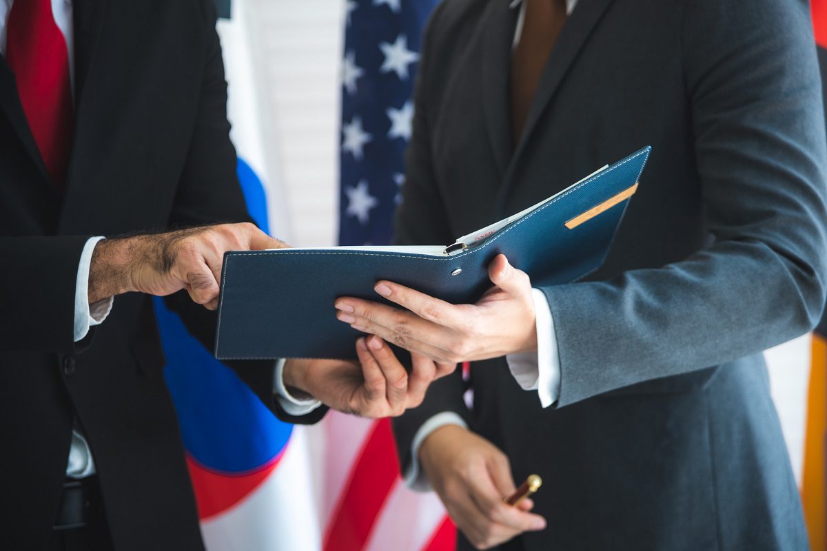 Two people in suits review a document folder, with the flags of the United States and South Korea visible in the background.