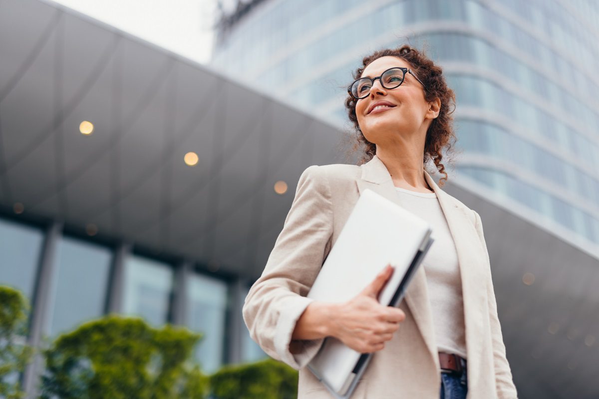Woman wearing glasses and a beige blazer stands outside a modern glass building, holding a laptop and smiling.