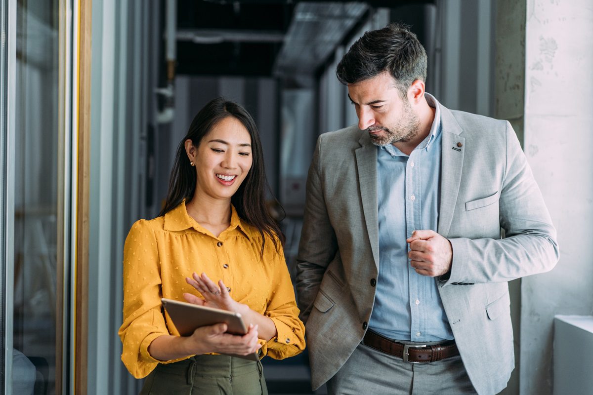 Shot of a two confident business persons talking in the work place. Two colleagues using a digital tablet while walking in a modern office. Businessman and businesswoman in meeting discussing business strategy. Business coworkers working together in the office.