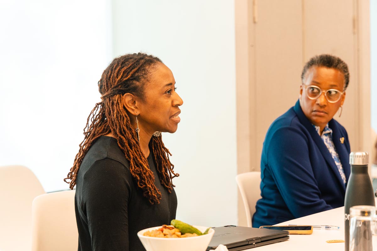 Two women sit at a table in a meeting room; one is speaking while the other listens. A laptop, a bowl of food, and a water bottle are on the table.