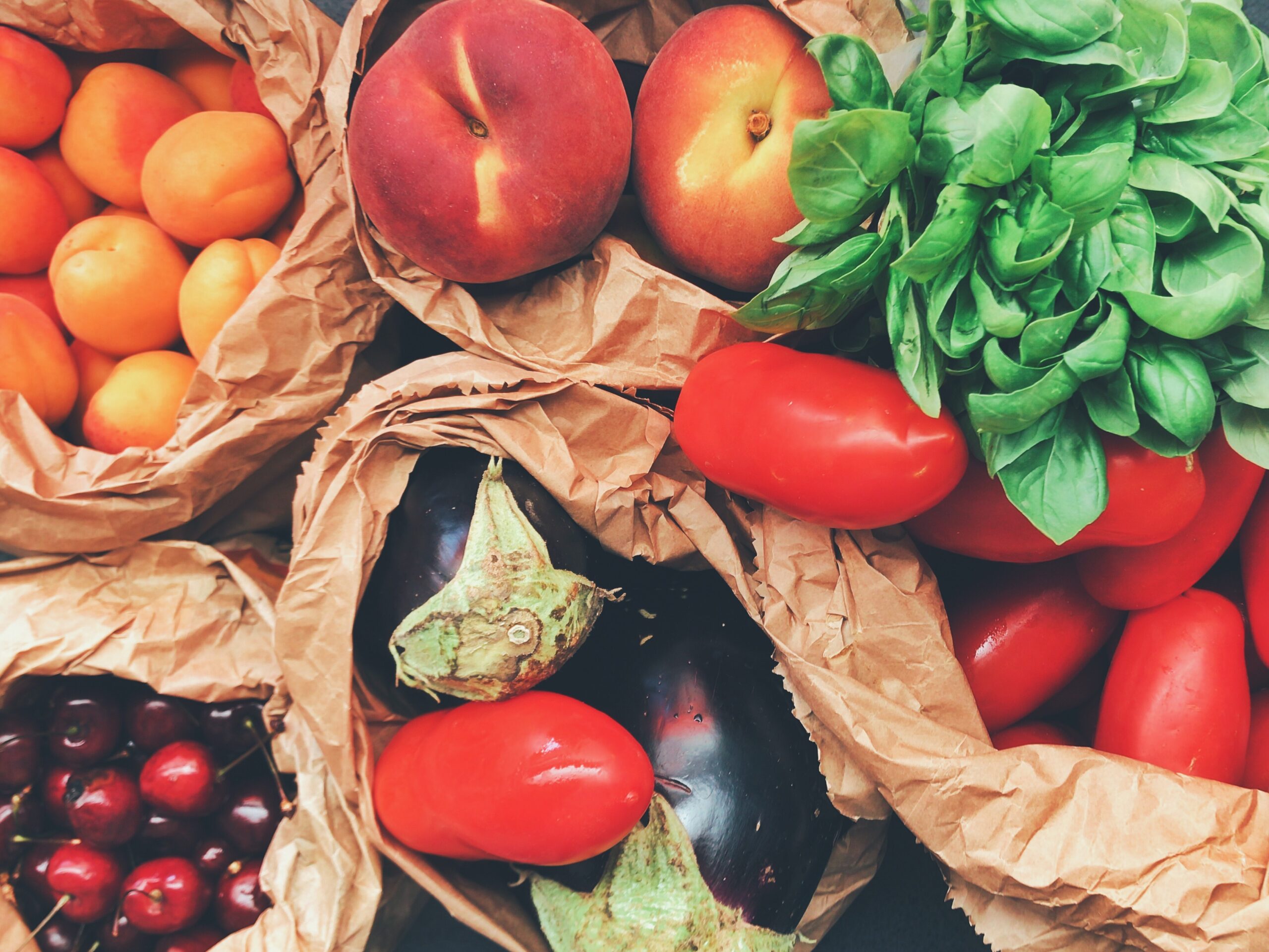Bags of fresh produce including apricots, peaches, basil, tomatoes, eggplants, and cherries arranged in brown paper wrappers.