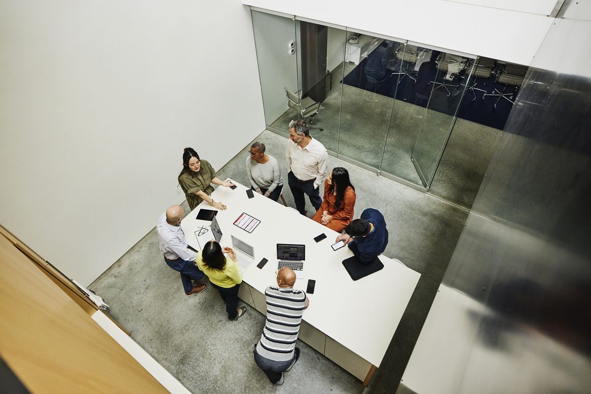 Seven people are meeting around a white table in a modern office, using laptops and tablets, viewed from above.