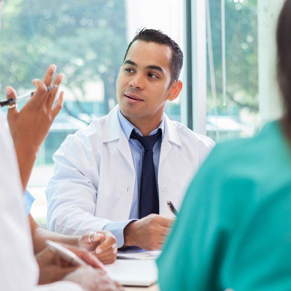 In a bright room, a male doctor in a white coat takes notes and looks at another person out of frame