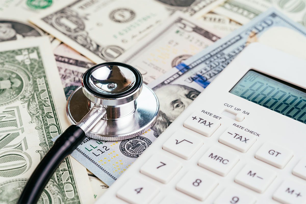 A stethoscope and a calculator rest on top of scattered US dollar bills, symbolizing the financial costs of healthcare.