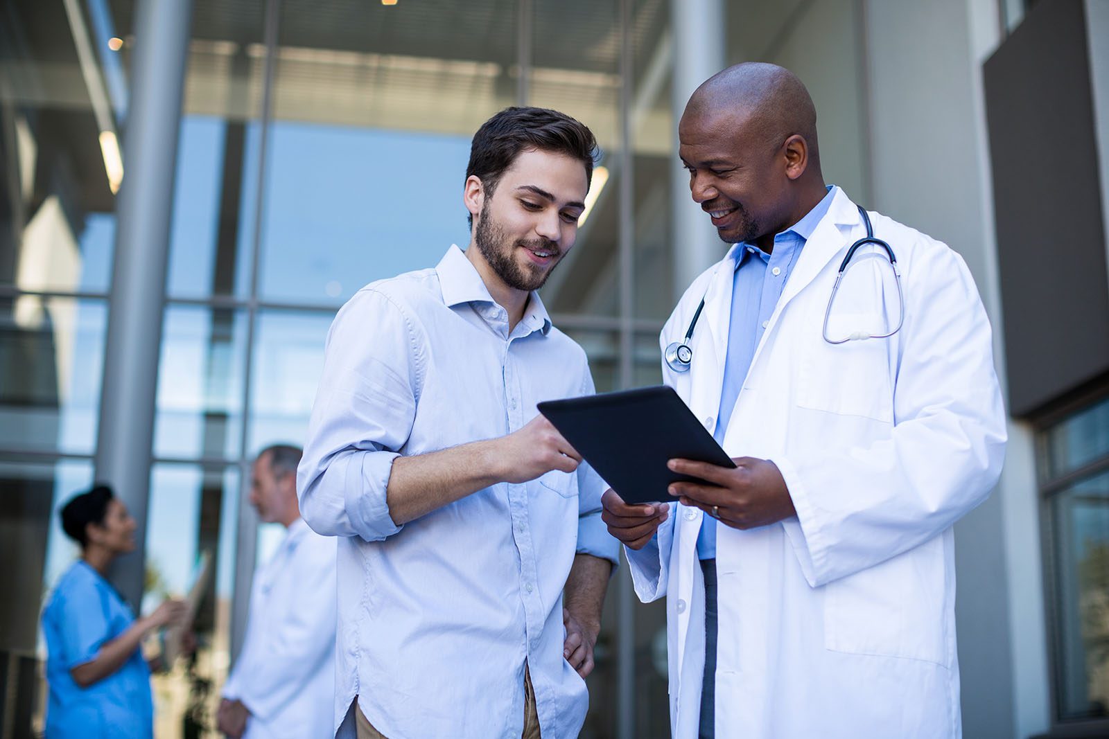A doctor in a white coat shows a tablet to a patient in a modern medical facility, while two other medical staff talk in the background.