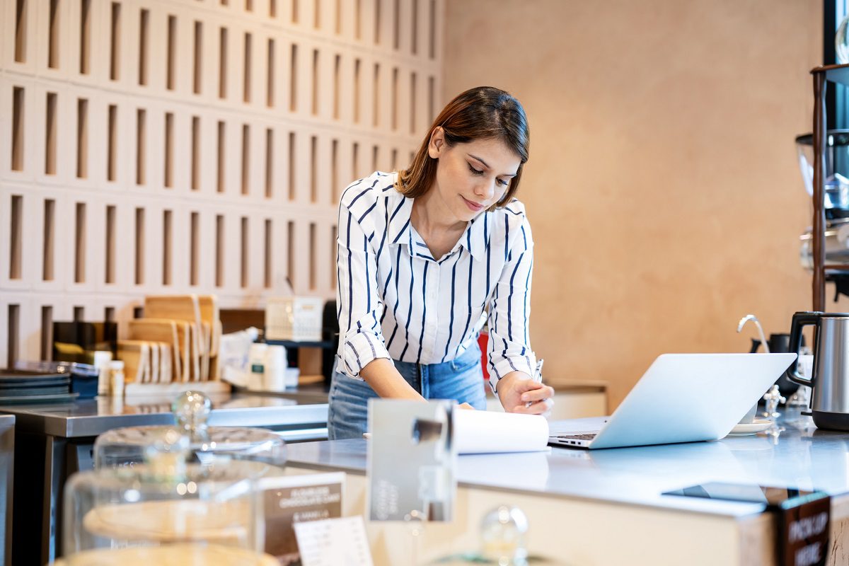 A woman stands at a counter in a modern cafe, writing on paper while looking at an open laptop.