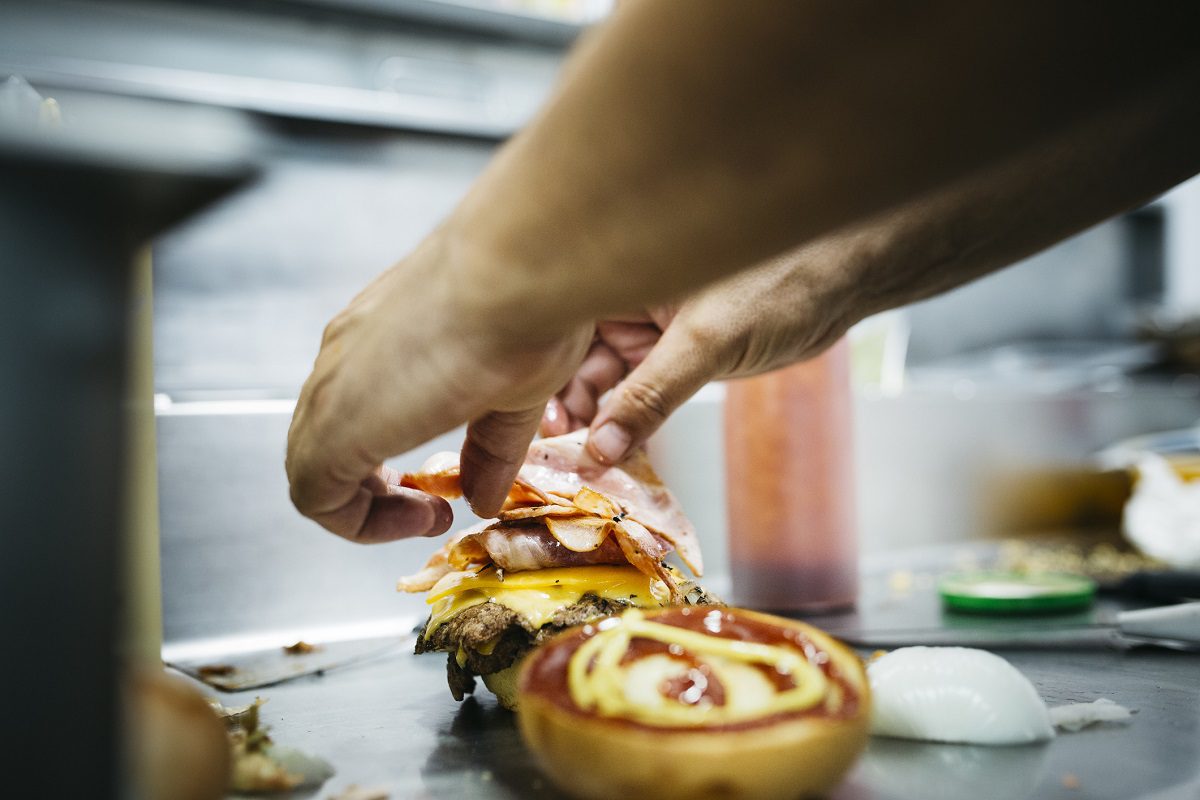 A person assembles a hamburger with layers of cheese, bacon, and ham, while a bun with ketchup and mustard sits nearby.
