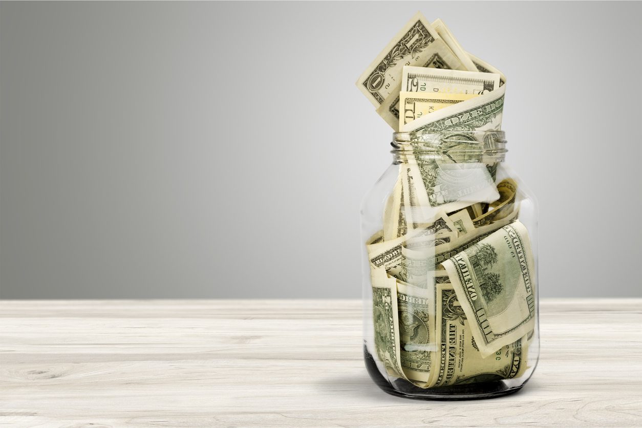 A glass jar filled with various denominations of U.S. dollar bills sits on a light wooden surface against a plain gray background.