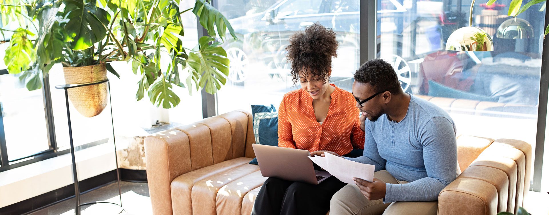 A man and woman discuss papers and point to a computer while sitting on a couch in front of big windows