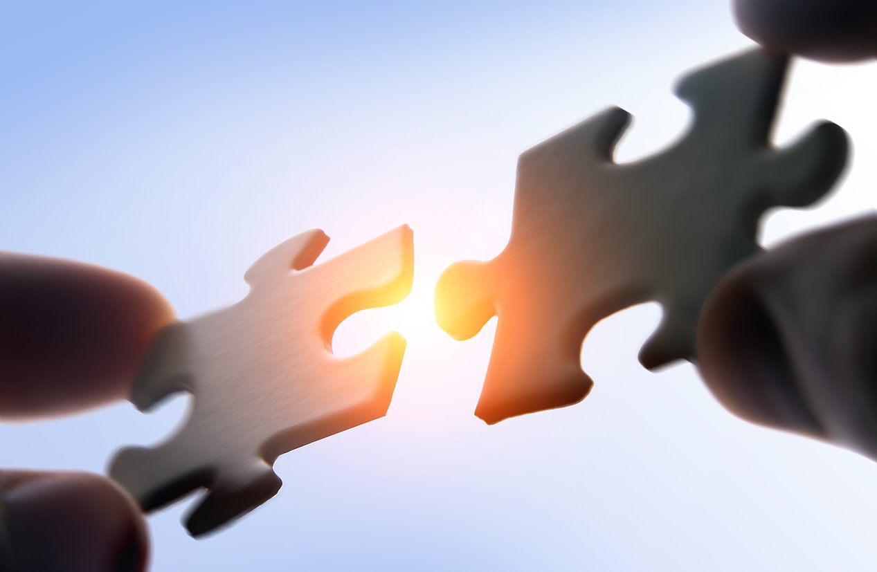 Two hands hold matching puzzle pieces close together with sunlight shining through the gap between them.