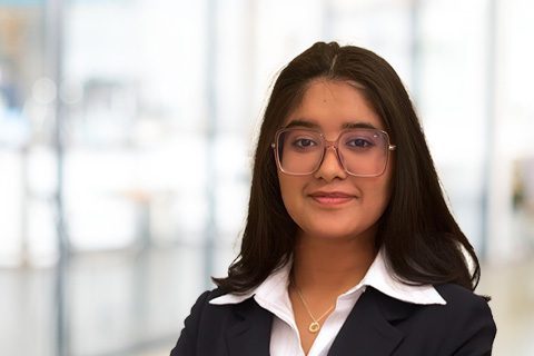 A young woman with long dark hair and glasses, dressed in a navy blazer and white shirt, stands in a bright, blurred office setting.