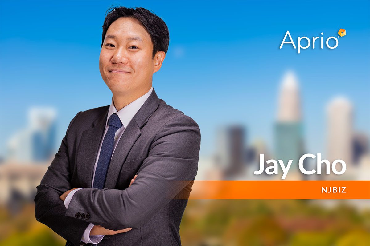 A man in a suit stands with arms crossed, smiling, in front of a blurred cityscape background. Text on the image reads Aprio, Jay Cho, and NJBIZ.