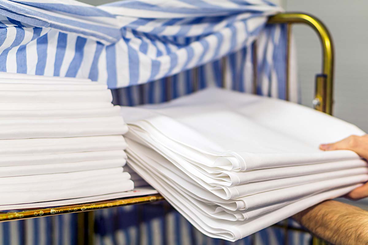 A person’s hands arranging neatly folded white linens on a cart, with a blue and white striped curtain partially visible in the background.