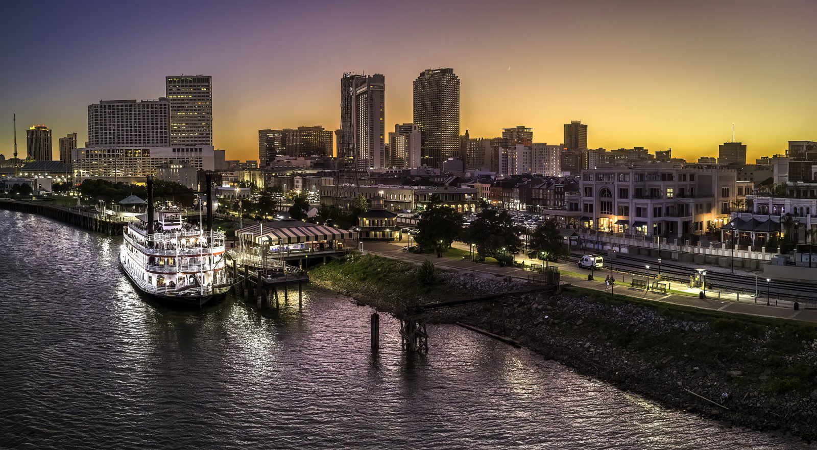 A riverboat is docked along the waterfront at sunset with a city skyline in the background and lights reflecting on the water.