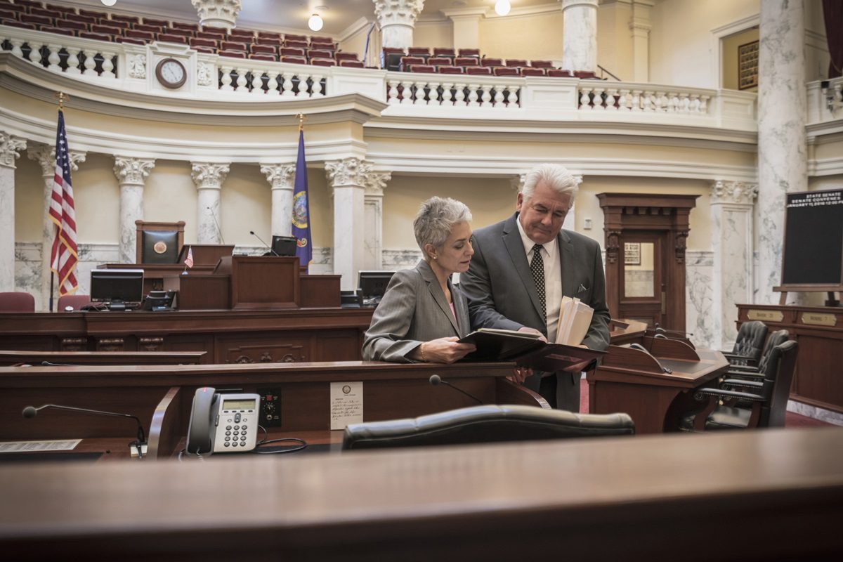 Two people in formal attire review documents together inside a large, empty courtroom with marble columns and flags.