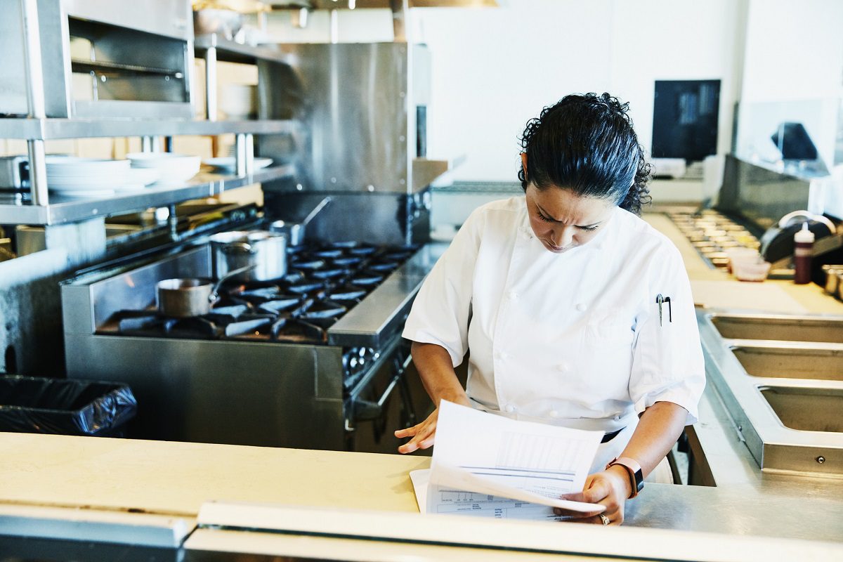 Chef in a white uniform reviews papers at a counter in a commercial kitchen with stoves, shelves, and cooking equipment in the background.
