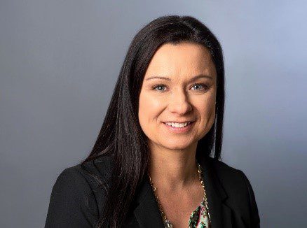A woman with long dark hair wearing a black blazer and a patterned top, smiling in front of a plain gray background.