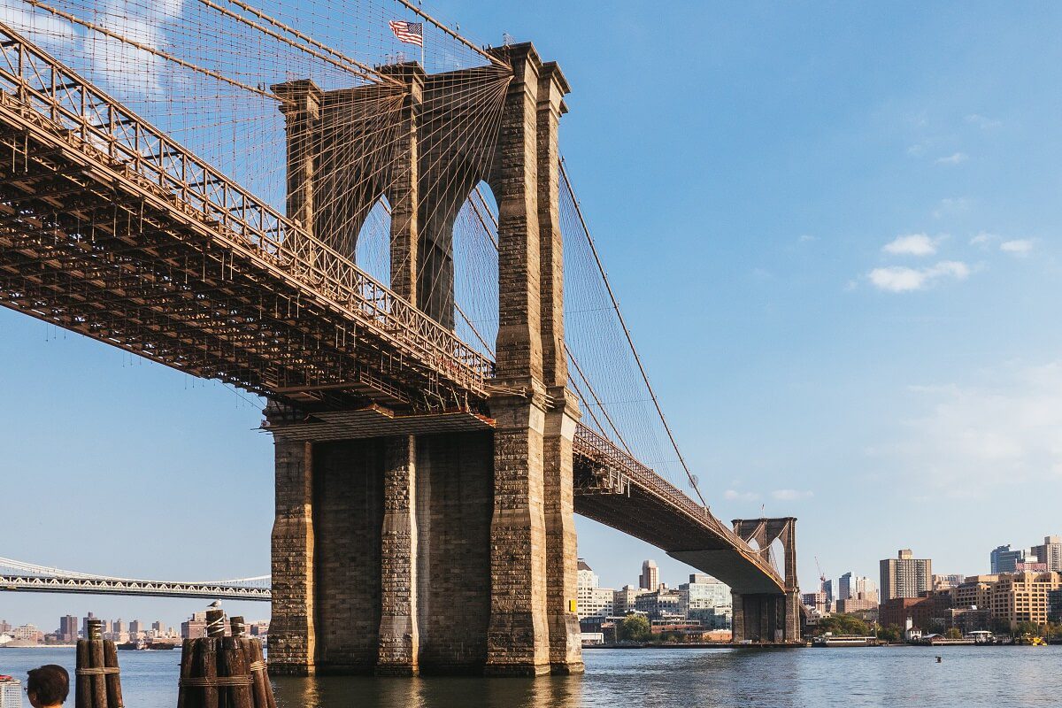 The Brooklyn Bridge spans the East River between Manhattan and Brooklyn under a clear sky, with city buildings visible in the background.