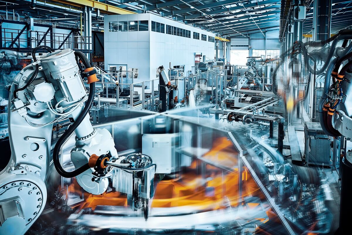 Robotic arms operate on an automated assembly line in a modern industrial factory, with a worker overseeing the machinery in the background.