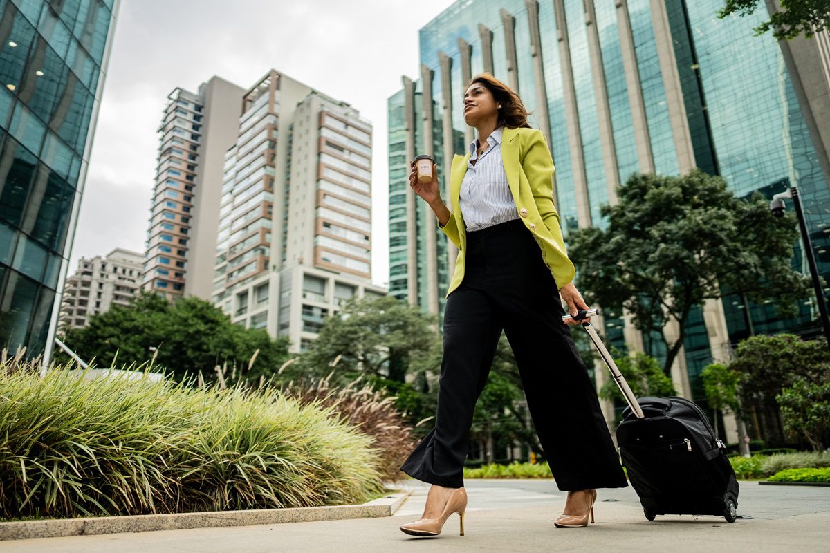A woman in business attire walks outdoors in a city with a suitcase and coffee cup, surrounded by tall buildings and greenery.