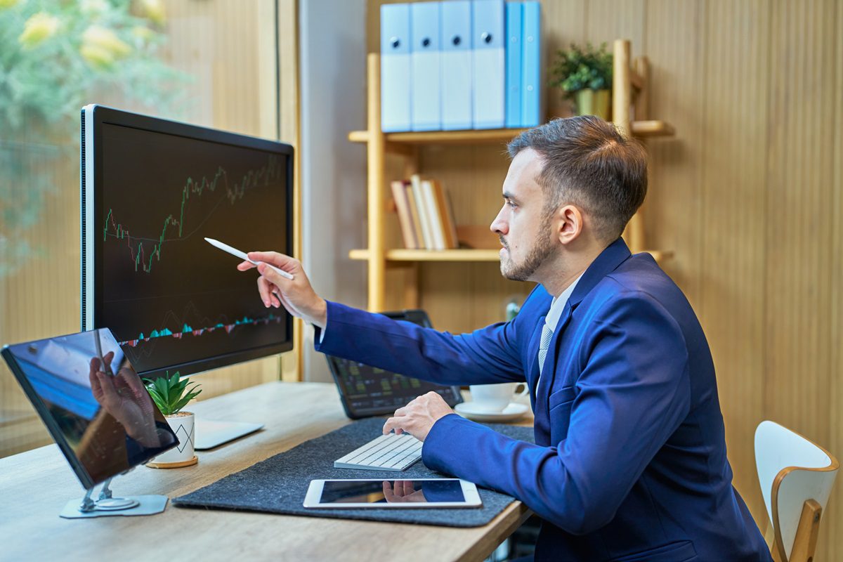 Young stockbroker pointing at a rising stock market graph displayed on a computer screen, analyzing financial data in a sleek, modern office environment