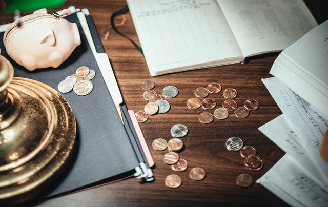 Scattered coins, a pink piggy bank, an open notebook, and papers are arranged on a wooden desk.