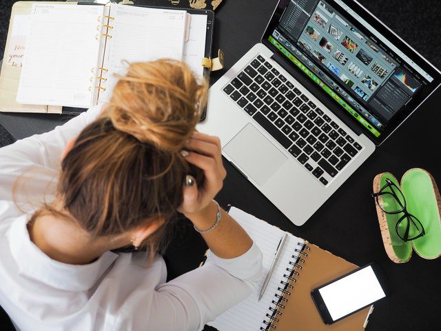 A person with their head in their hands sits at a desk with a laptop, notebooks, a phone, and glasses, appearing stressed while working.