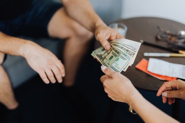 A person hands over a stack of U.S. dollar bills to another person sitting nearby at a table with papers, a pair of glasses, and a drink.