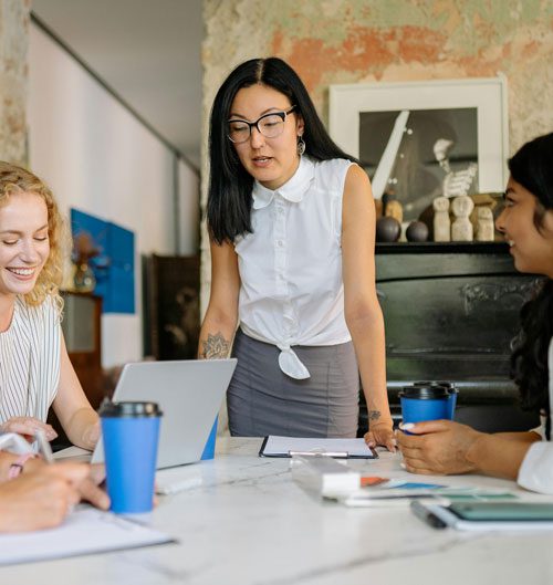 In an office setting, a businesswoman stands at the end of a conference room table and talks to employees