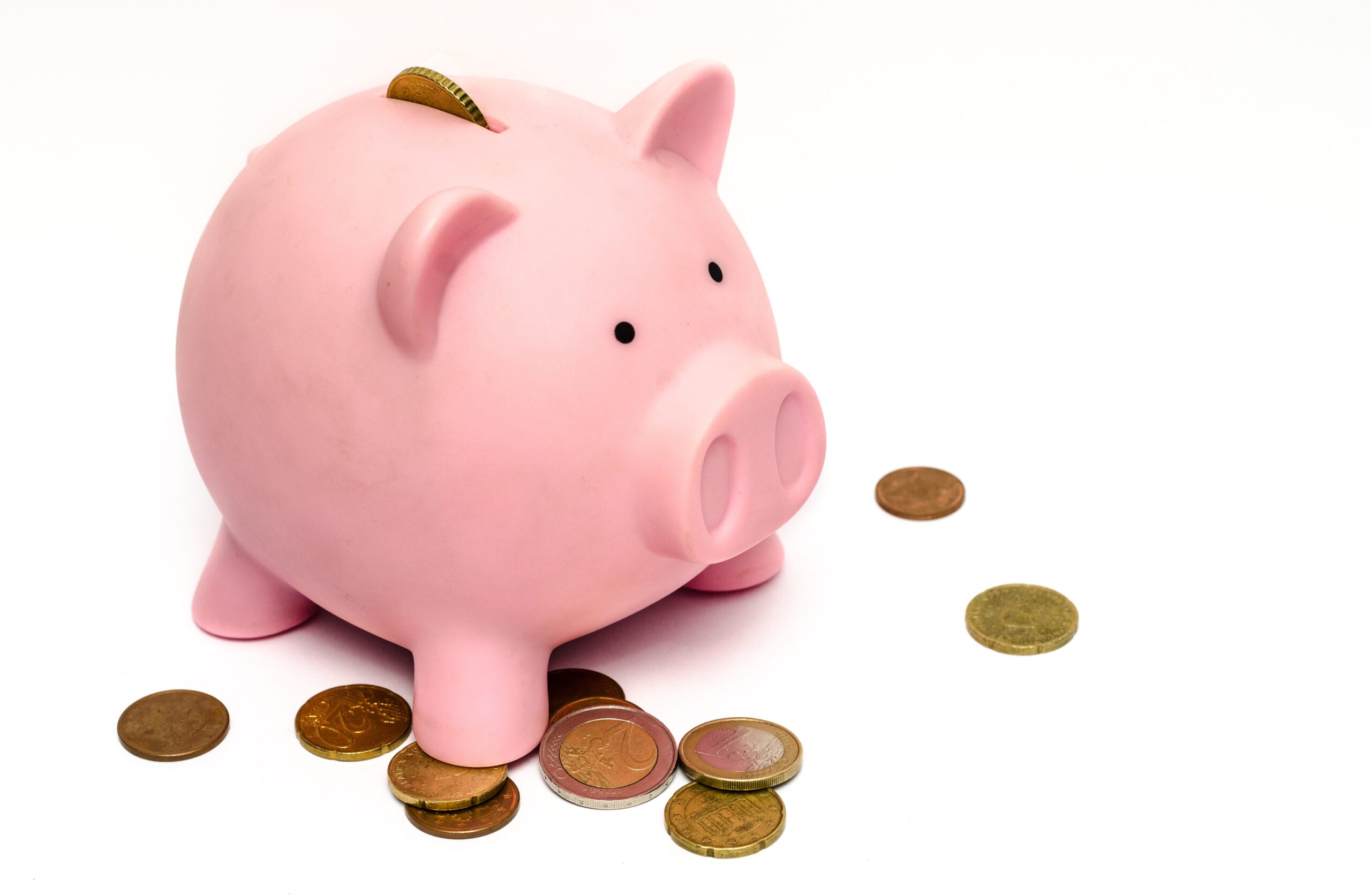 A pink piggy bank with coins scattered around it and one coin partially inserted in the slot on its back.