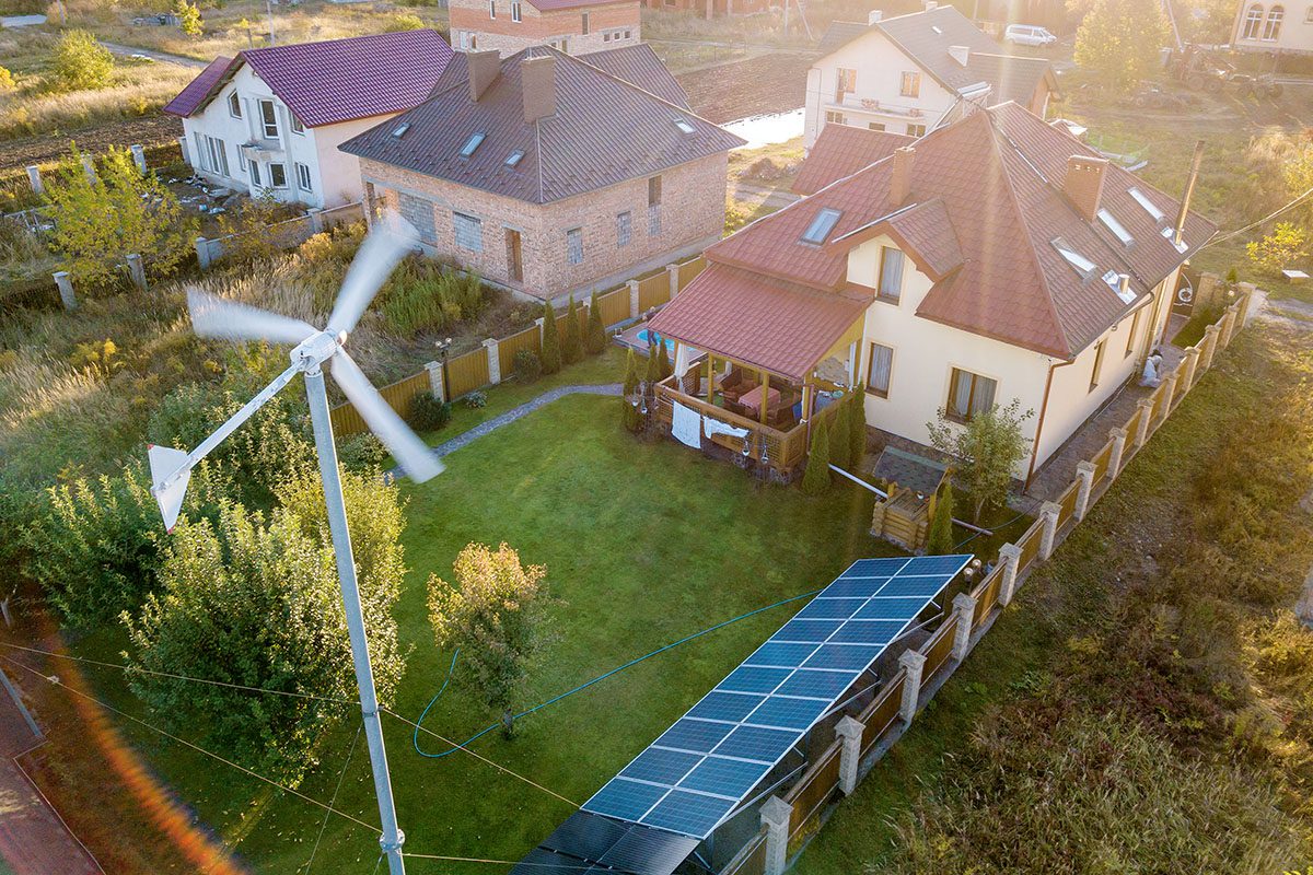 Aerial view of a house with solar panels on the lawn and a wind turbine in the yard, surrounded by a fence and neighboring houses.